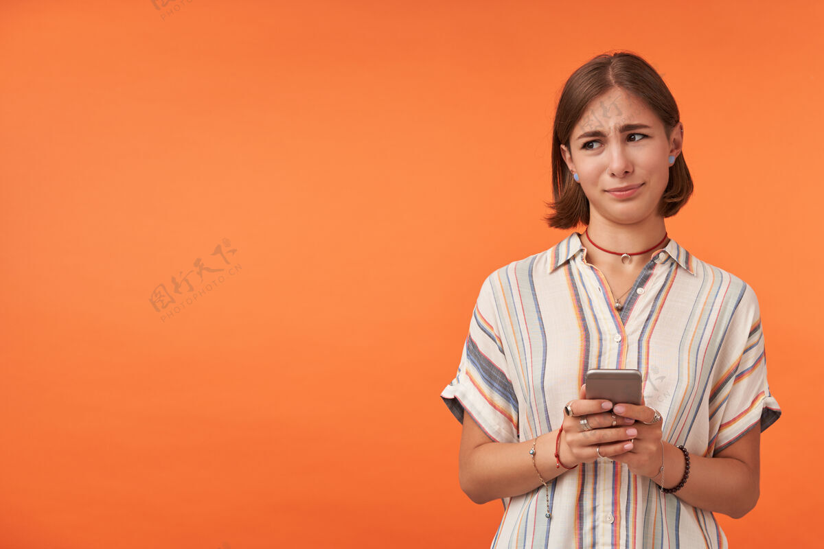 休闲漂亮的学生拿着一部智能手机 不相信她所看到的穿着条纹衬衫 戴着牙套和手镯看着左边靠着橙色墙的复印空间女性青少年社交