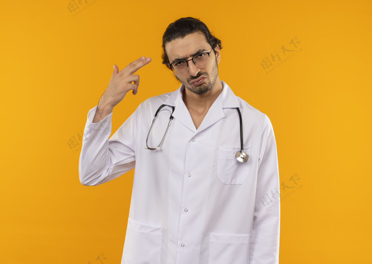 医生悲伤的年轻医生戴着医用眼镜 穿着医用长袍 听诊器在隔离的黄色墙上显示着手枪的手势 还有复印空间长袍眼镜年轻