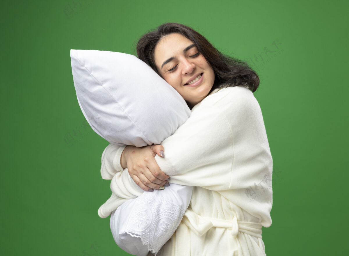 长袍微笑的年轻白种人患病女孩穿着长袍站在侧视图拥抱枕头闭上眼睛隔离在绿色背景女孩年轻枕头