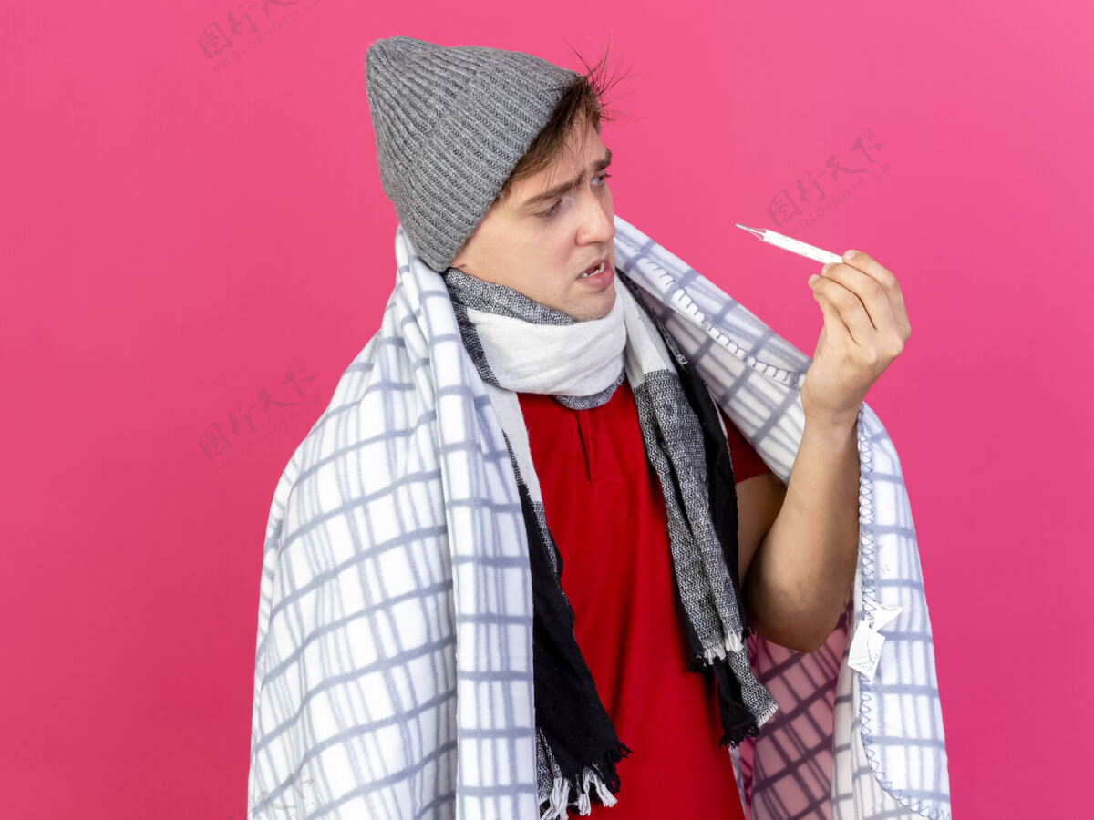 帅哥年轻英俊的金发病夫戴着冬天的帽子 围着格子呢围巾 手里拿着温度计 看着被隔离在深红色背景下的复制空间抱着围巾裹着