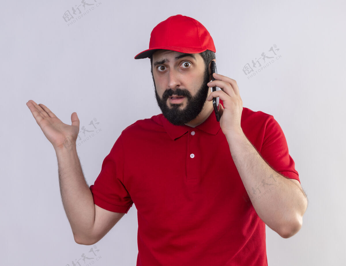 手印象深刻的年轻英俊的白人送货员穿着红色制服和帽子讲电话 并显示空手孤立在白色送货帽子年轻