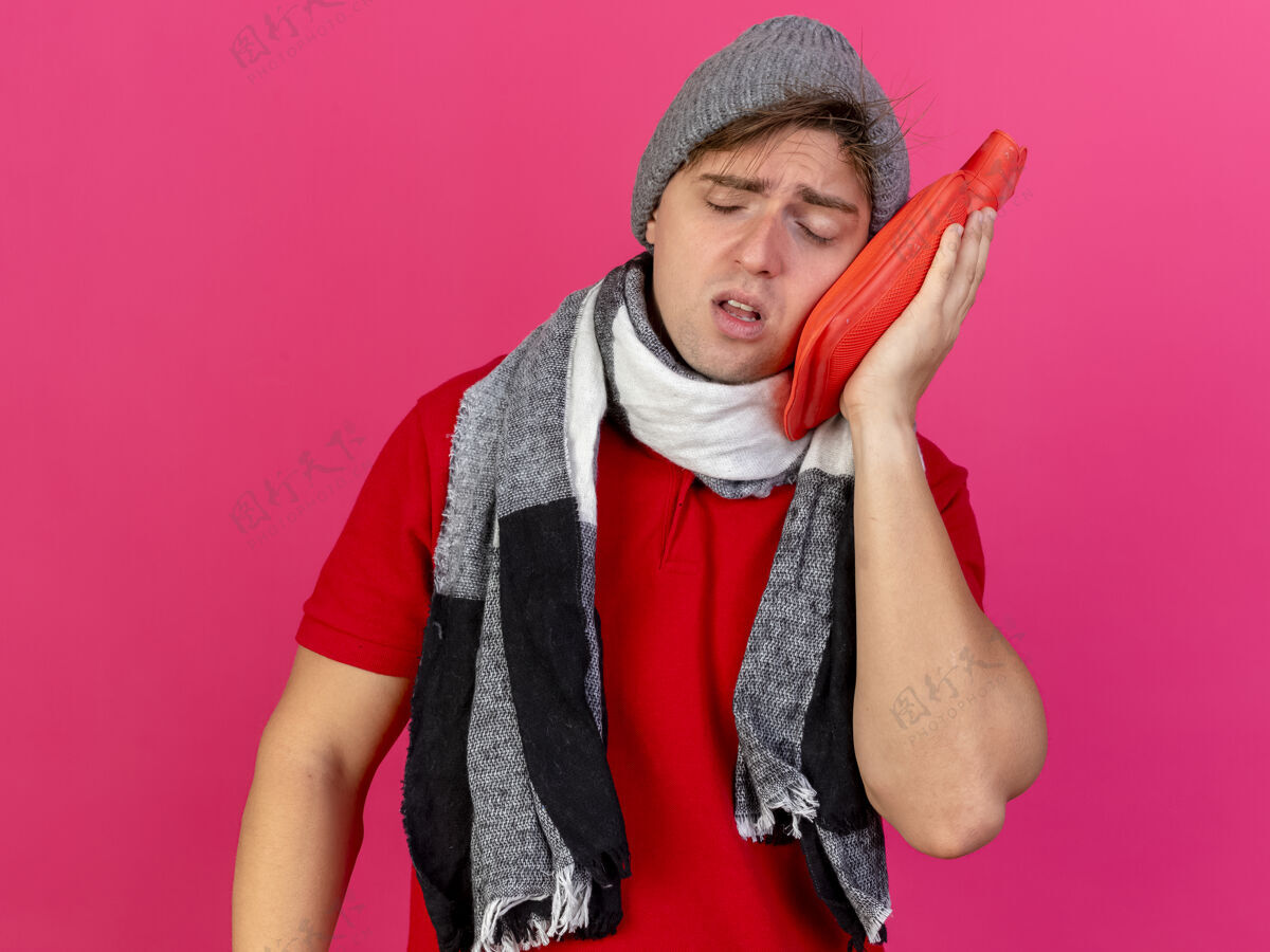 年轻瘦弱的年轻英俊的金发病夫戴着冬天的帽子和围巾拿着热水瓶在深红色的背景上闭着眼睛抚摸着脸疾病帽子触摸