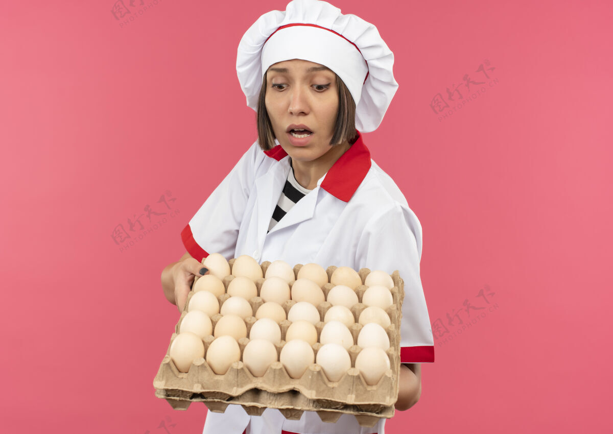 印象印象深刻的年轻女厨师穿着厨师制服 拿着一盒鸡蛋 看着粉红色的鸡蛋与复印空间隔离开来持有年轻空间