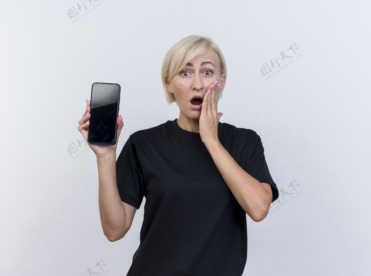 脸颊惊讶的中年金发斯拉夫妇女展示手机 手放在脸颊上 看着隔离在白色背景上的相机 还有复印空间惊喜手机电话