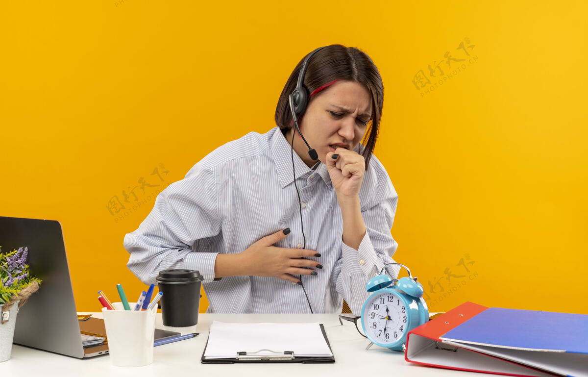 办公桌生病的年轻呼叫中心女孩戴着耳机坐在办公桌旁咳嗽 手放在胸前 孤立在橘子上电话咳嗽耳机
