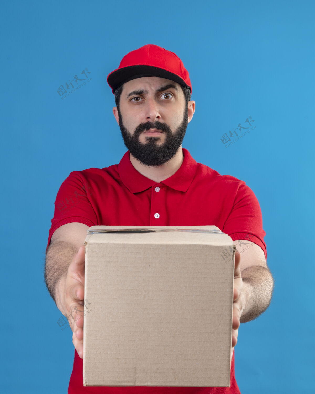 迷糊困惑的年轻英俊的白人送货员穿着红色制服 戴着帽子 伸出纸箱对着蓝色的摄像机年轻制服男人