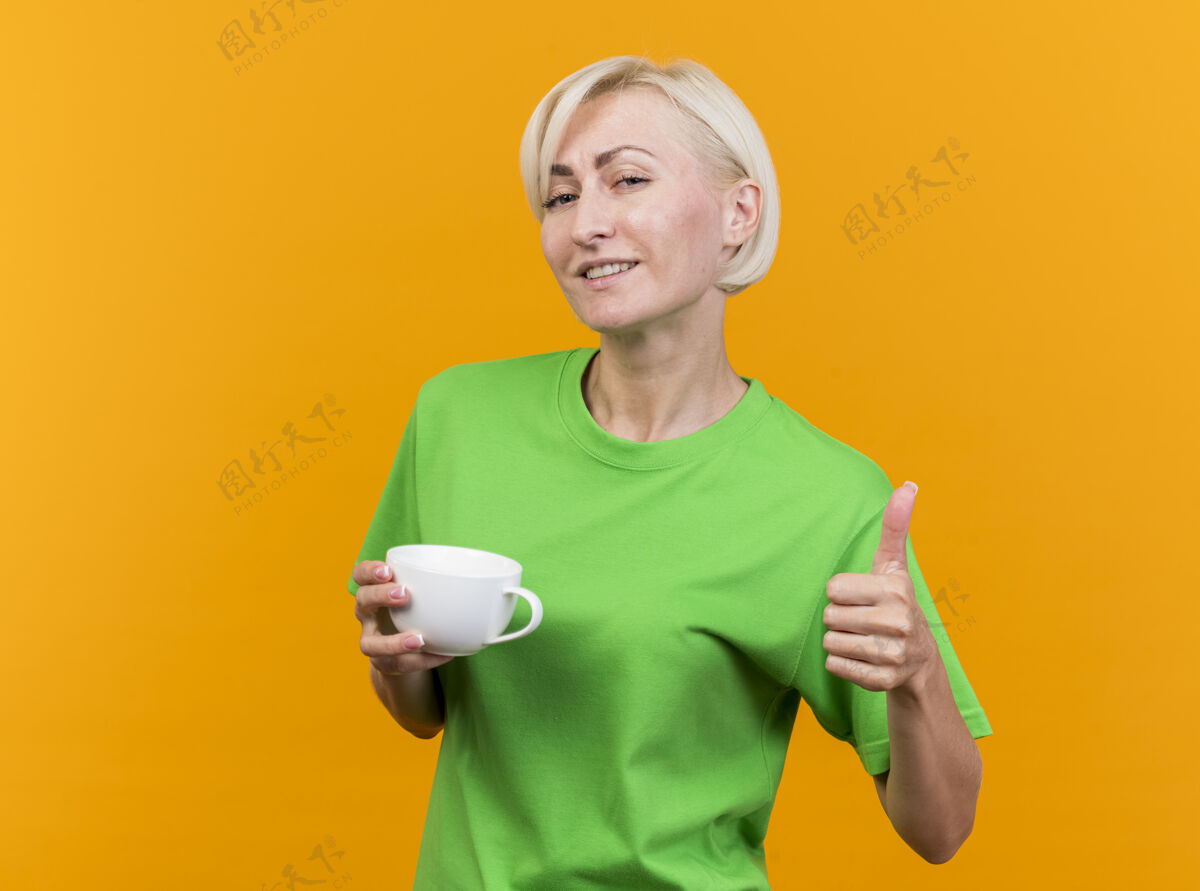 茶高兴的中年金发斯拉夫妇女拿着一杯茶看着相机显示拇指向上隔离在黄色背景与复制空间拿着杯子金发