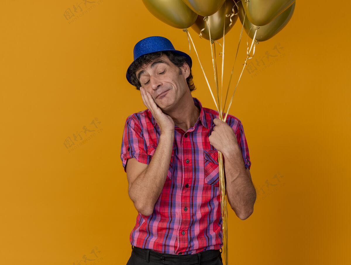 脸高兴的中年白种人党男子戴党帽举行气球触摸脸与封闭的眼睛隔离在橙色背景与复制空间抱着眼睛中年人