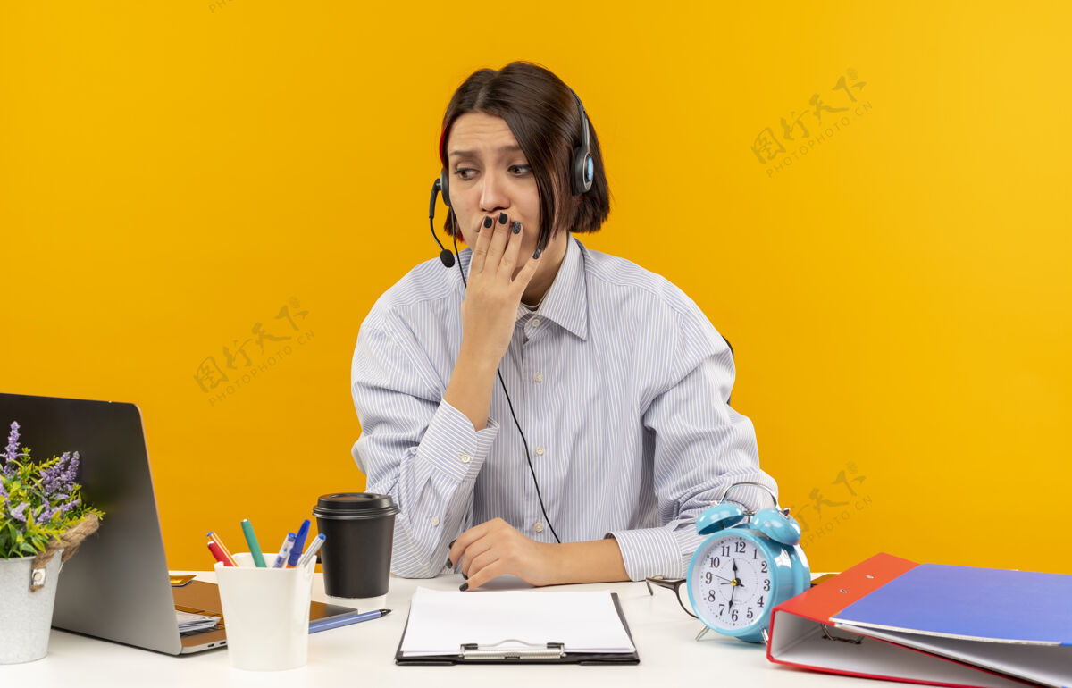 办公桌焦急的年轻呼叫中心女孩戴着耳机坐在办公桌旁 手放在嘴边看着橙色的隔离物手女孩坐着