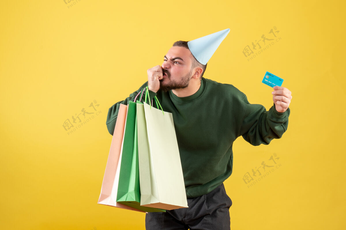 圣诞节正面图：年轻男性手持黄色背景的银行卡和购物包帽子聚会男性