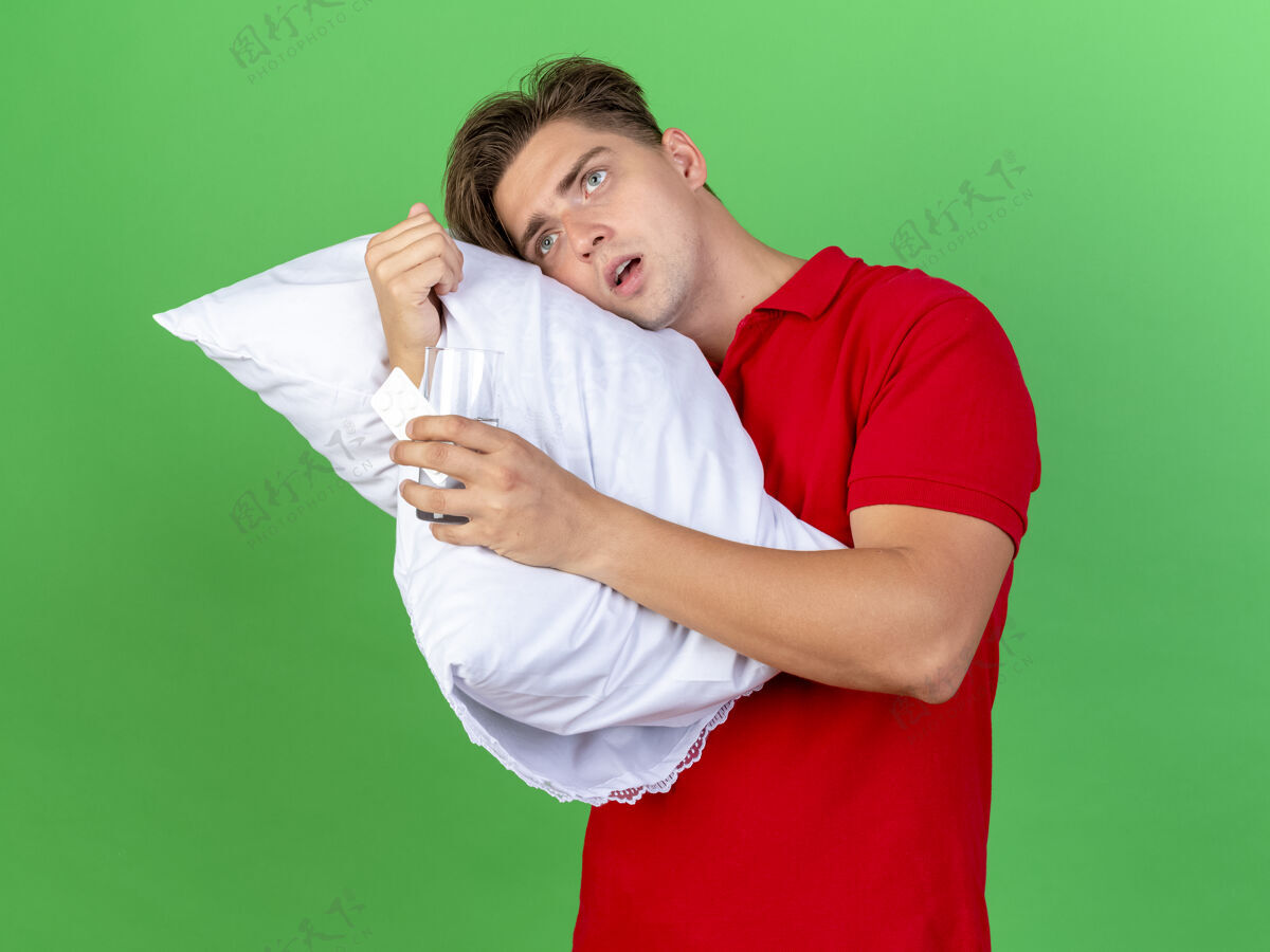 印象印象深刻的年轻英俊的金发病男子抱着枕头把头放在上面拿着一包药片看着绿色背景上孤立的一面英俊金发持有