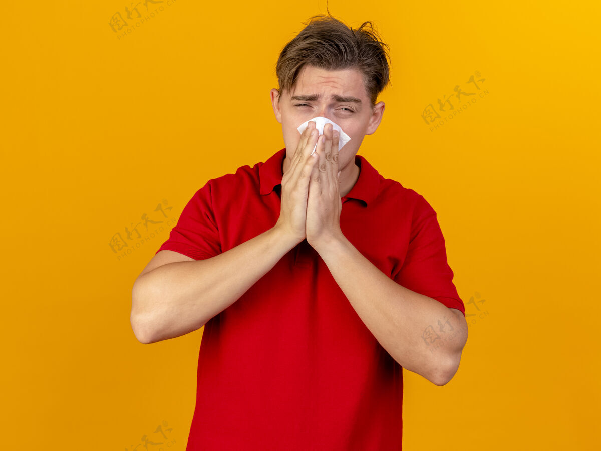 年轻年轻英俊的金发病夫看着相机擦鼻子用餐巾隔离在橙色背景与复制空间金发疾病鼻子