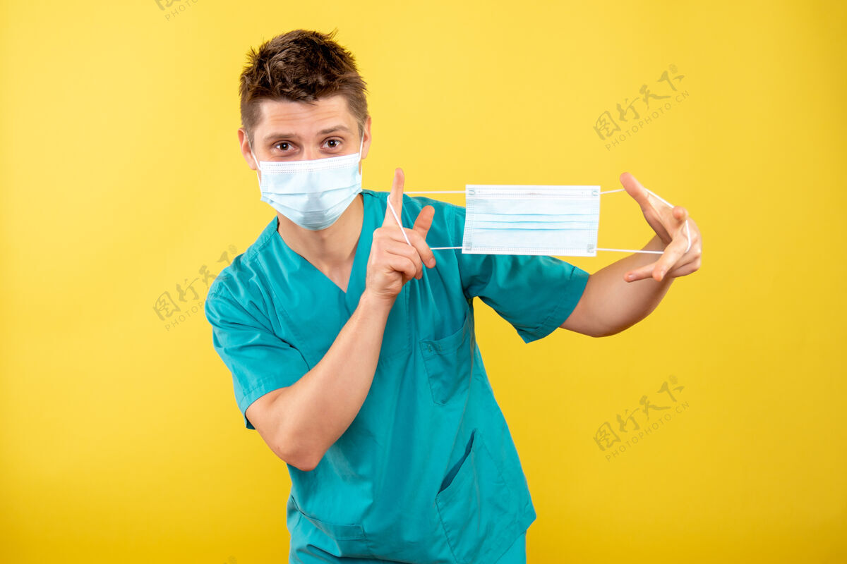 医学正面图年轻的男医生穿着医疗服 戴着无菌口罩 拿着黄色背景上的另一个口罩人人背景