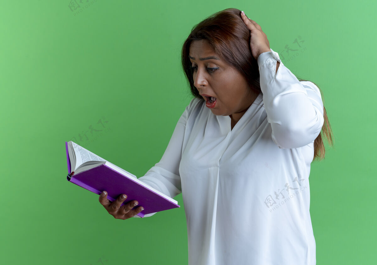 抱惊讶的是 随意的白人中年妇女抱着书 看着书 把手放在隔离在绿色墙上的头上头休闲女人