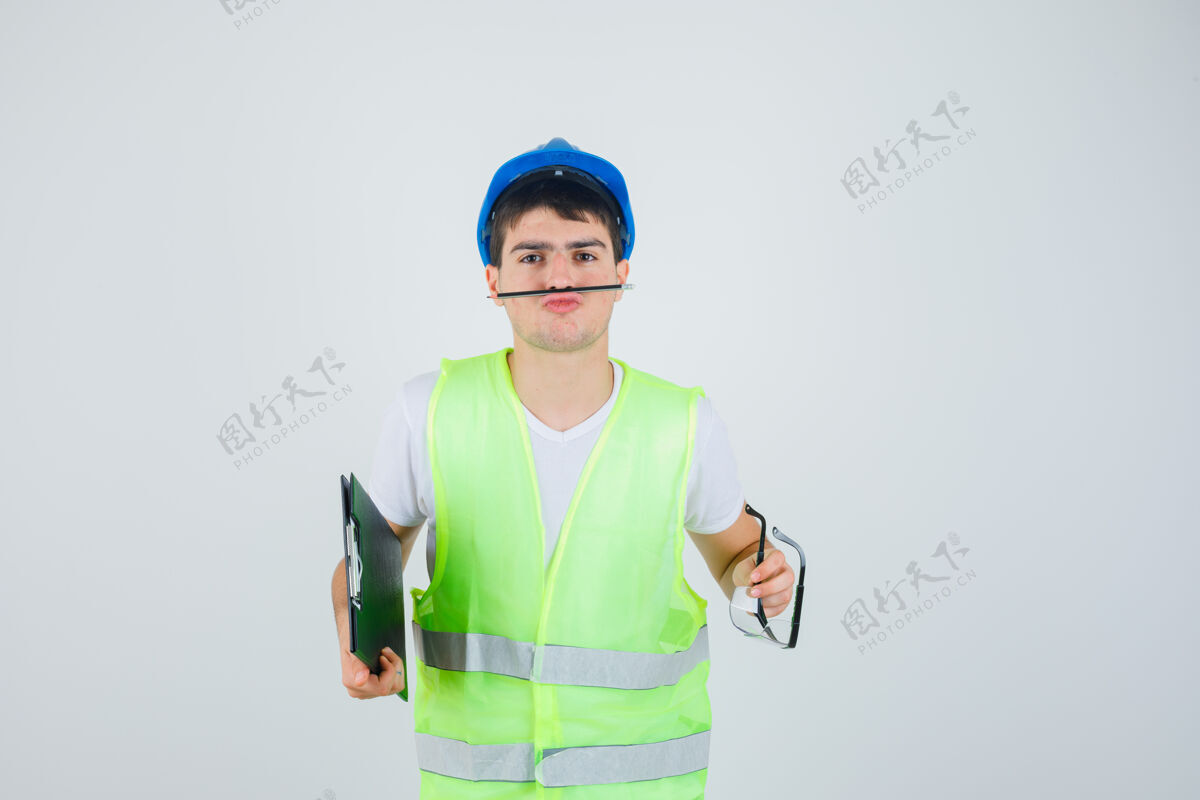 表情穿着建筑制服的小男孩手里拿着剪贴板和安全眼镜 嘴里拿着钢笔 神情严肃 前视建筑人眼睛