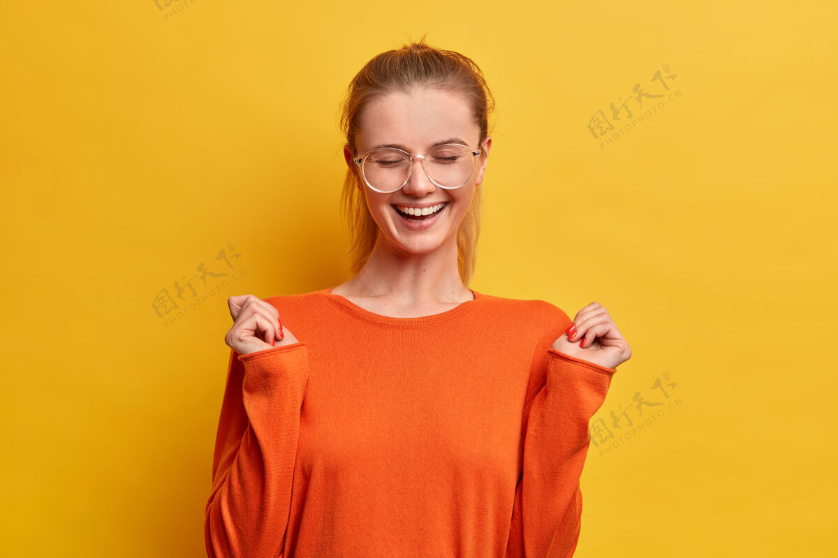 心情快乐乐观的女孩 带着快乐的微笑 闭上眼睛 举手 对着积极的事情咯咯笑 戴着眼镜和橙色套头衫 肖像透明眼镜
