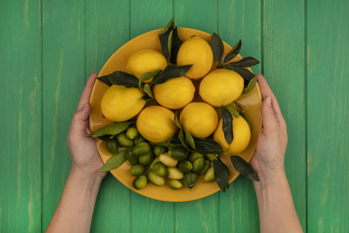 素食者顶视图中的女性手拿着一桶新鲜水果 如柠檬和金盏花在绿色的木墙上木材饮食农业