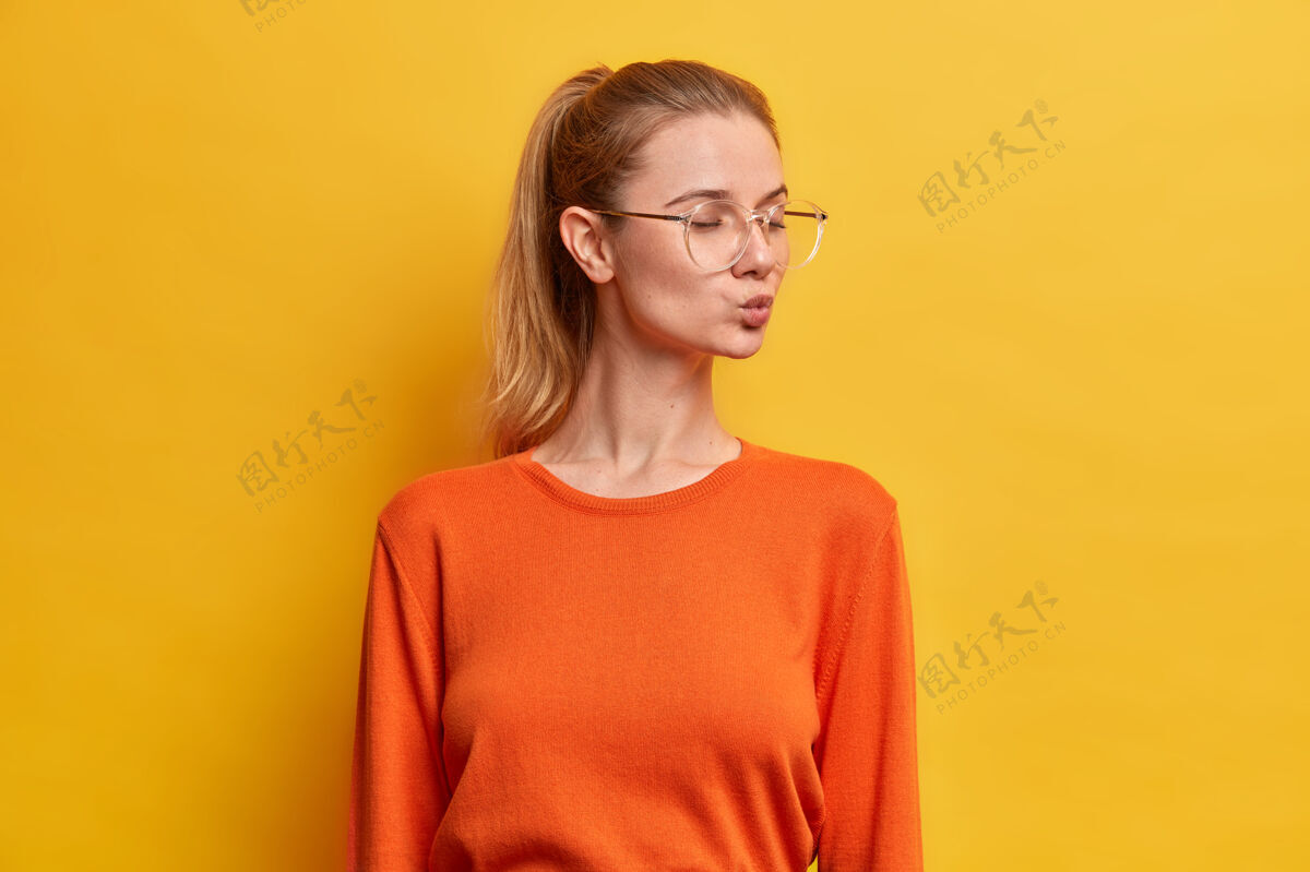 眼镜美丽的欧洲年轻女子闭着眼睛站着 嘴唇圆润 有着浪漫的心情 马尾辫 穿着橙色的套头衫 转身漂亮套头衫