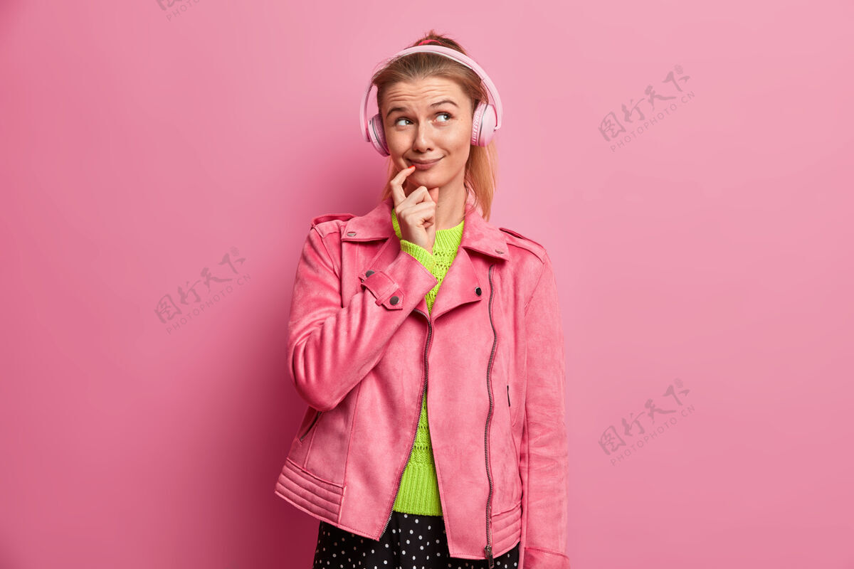 娱乐微笑迷人的千禧一代女性 表情梦幻 聆听喜爱的歌曲 戴着耳机 欣赏播放列表 穿着粉色夹克 站在室内爱好 休闲 生活方式声音快乐女性
