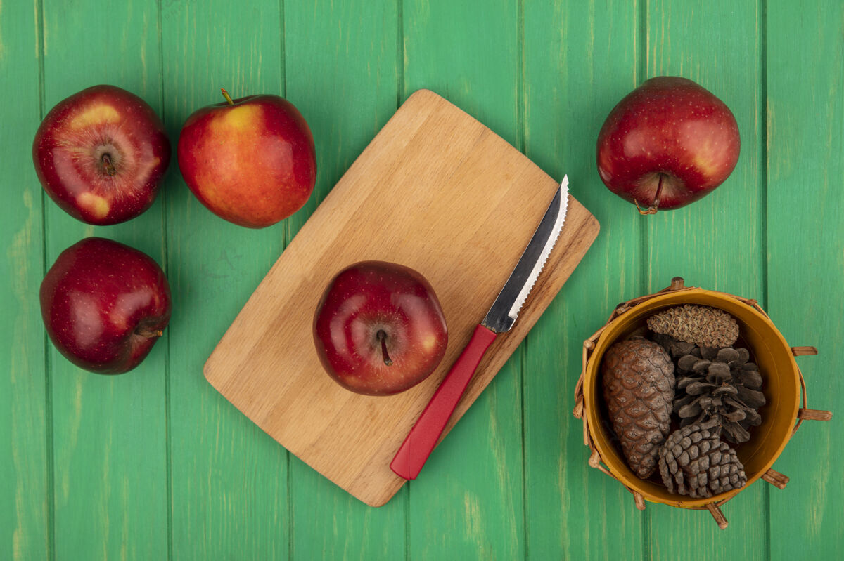 视图一个健康的红苹果在木制厨房板上的俯视图 刀子上放着松果 桶里的苹果被隔离在绿色的木墙上厨房水果桶