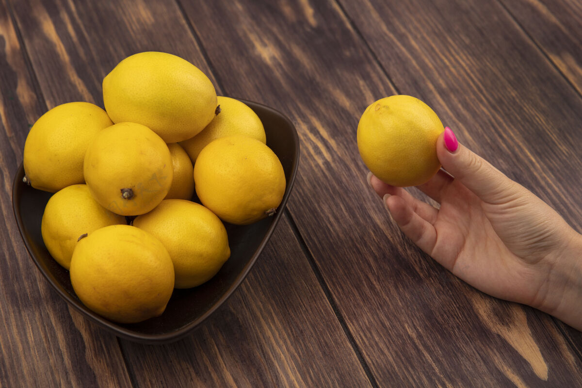 块顶视图中的女性手拿着一个新鲜的柠檬和一碗柠檬在木墙上水果碗顶部