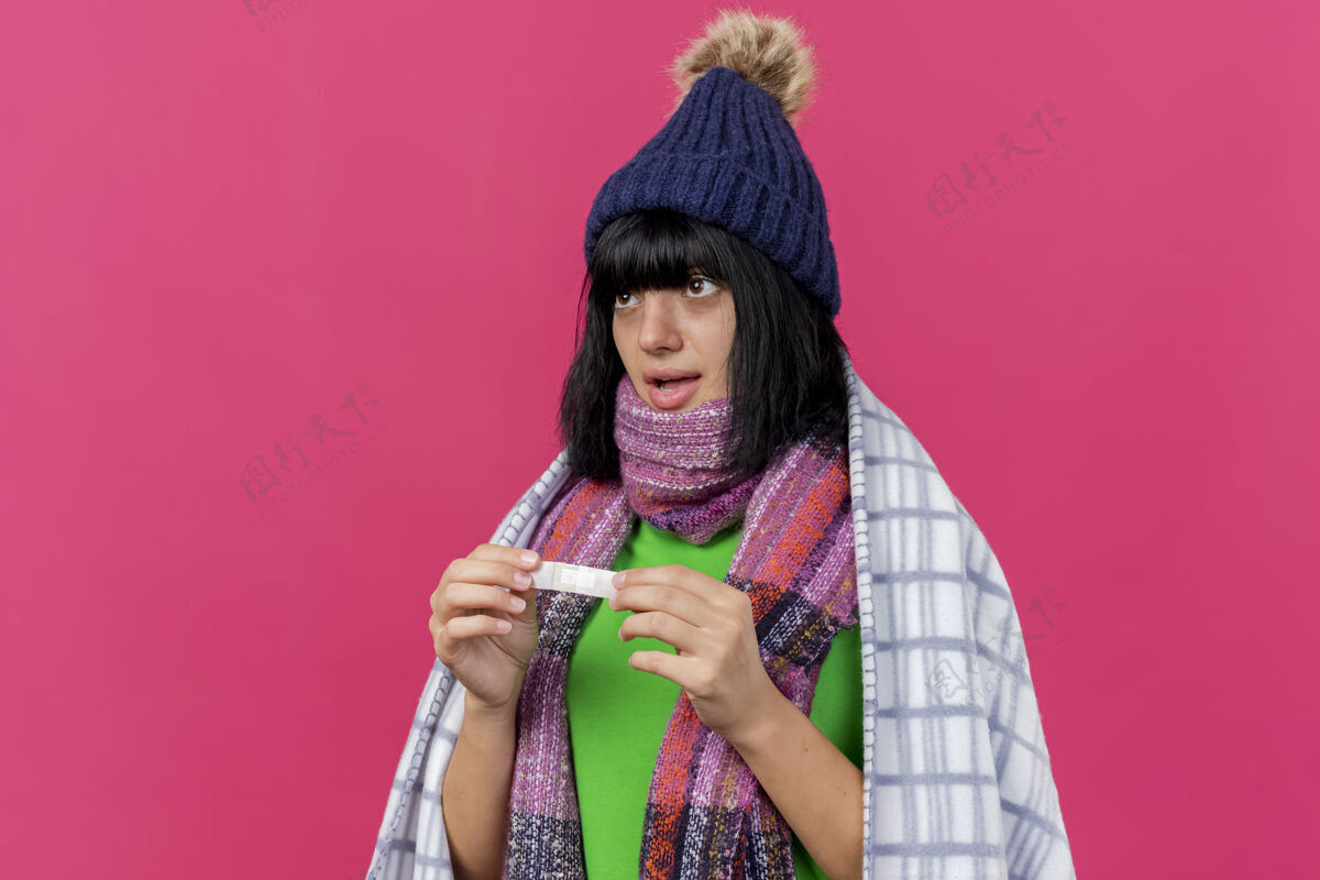 直令人印象深刻的年轻病高加索女孩戴着冬季帽子和围巾包裹在格子布举行医疗石膏看直隔离在深红色背景与复制空间穿格子深红色