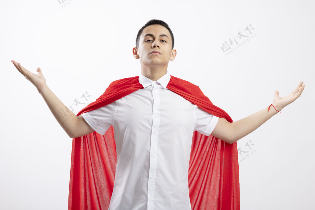 信心穿着红色斗篷的自信的年轻超级英雄男孩看着镜头 双手分开放在白色背景上超级英雄蔓延男孩