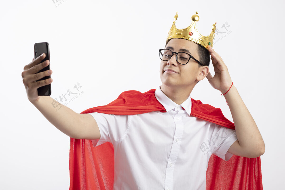 红色穿着红色斗篷戴着眼镜和皇冠的年轻超级英雄男孩在白色背景下独自自拍眼镜年轻男孩