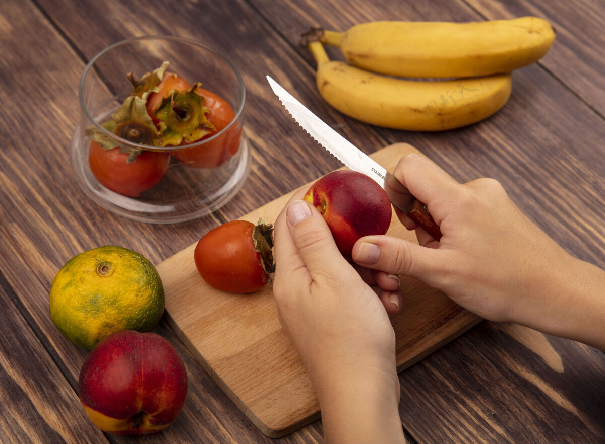 厨房俯视图中的女性手切割一个多汁的桃子在一个木制的厨房板上用刀与橘子和香蕉隔离在一个木制的墙壁木材桃有机