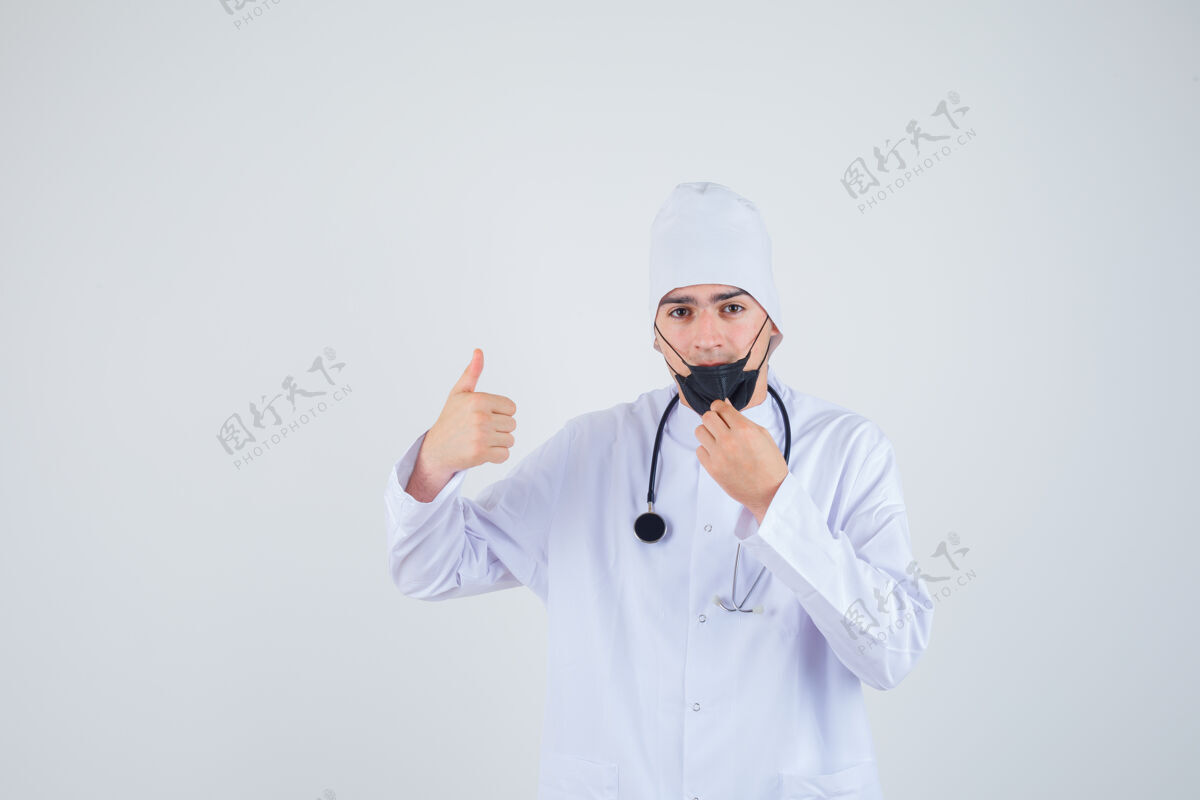 年轻年轻人试着拿起口罩 穿着白色制服 戴着口罩 大拇指朝上 看上去很满意正视图信心男性医生