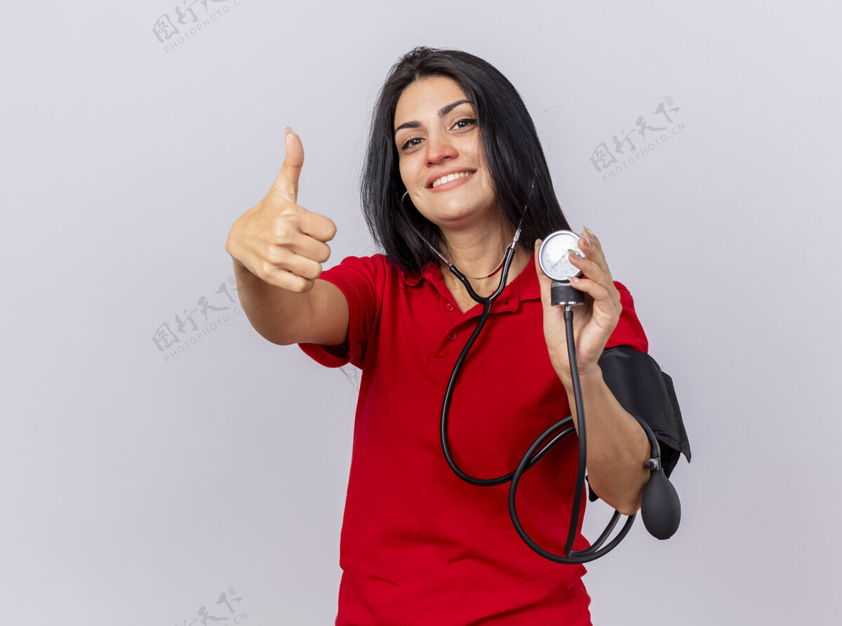 佩戴带着听诊器的年轻白种人女孩微笑着看着摄像机 用血压计测量她的血压 在白色背景上孤立地竖起大拇指 留有复印空间年轻压力血压计