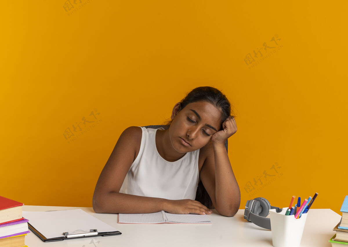 手疲惫的年轻女学生闭着眼睛坐在课桌旁 手里拿着学习用具 头放在桔子上桌子坐着桔子