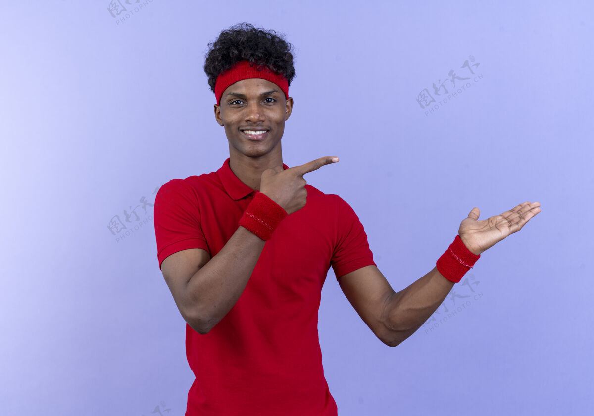 腕带高兴的年轻黑人美国运动男子戴着头带和腕带点与手和手指对侧隔离在蓝色墙上的复制空间穿壁板手指