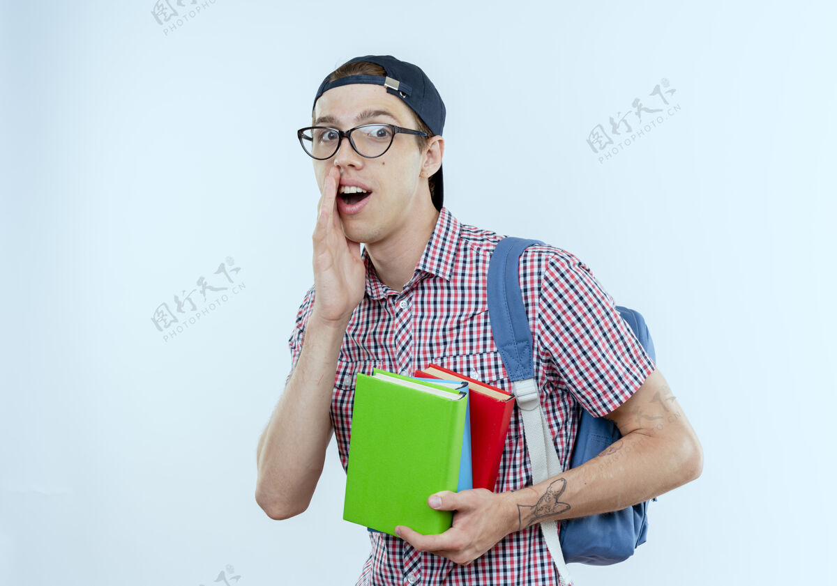 包高兴的年轻学生男孩戴着背包 眼镜和帽子拿着书和白色的耳语学生拿着眼镜