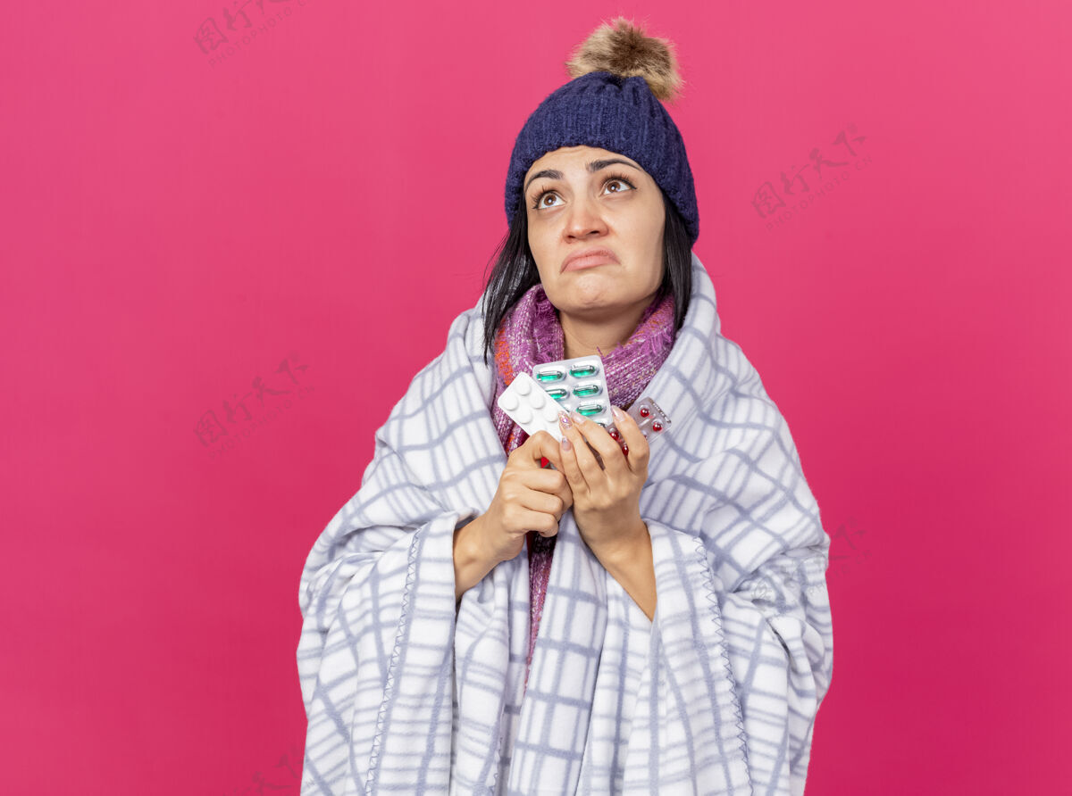 包装困惑的年轻生病的女人戴着冬天的帽子 围着方格布围巾 手里拿着一包包的药丸 孤零零地看着粉红色的墙上药丸帽子冬天