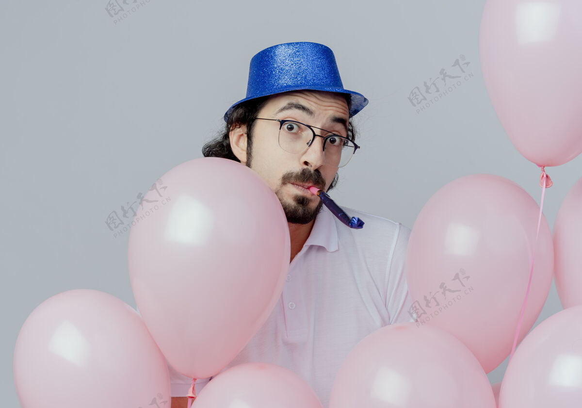 帅哥惊讶的帅哥戴着眼镜和蓝帽子站在气球后面 吹着白色的隔离哨子眼镜气球男人