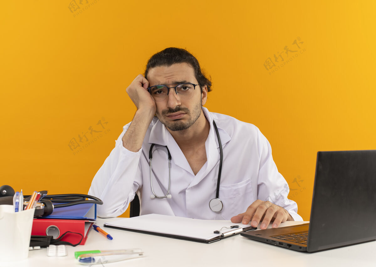 笔记本电脑悲哀的年轻男医生戴着医用眼镜穿着医用长袍带着听诊器坐着医疗年轻工作