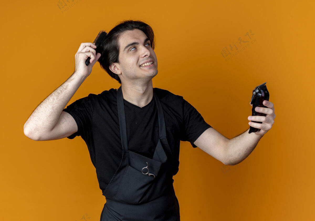 剪子年轻帅气的男理发师面带微笑 身着制服 手持理发器 梳着孤立的橙色头发橙色制服梳子