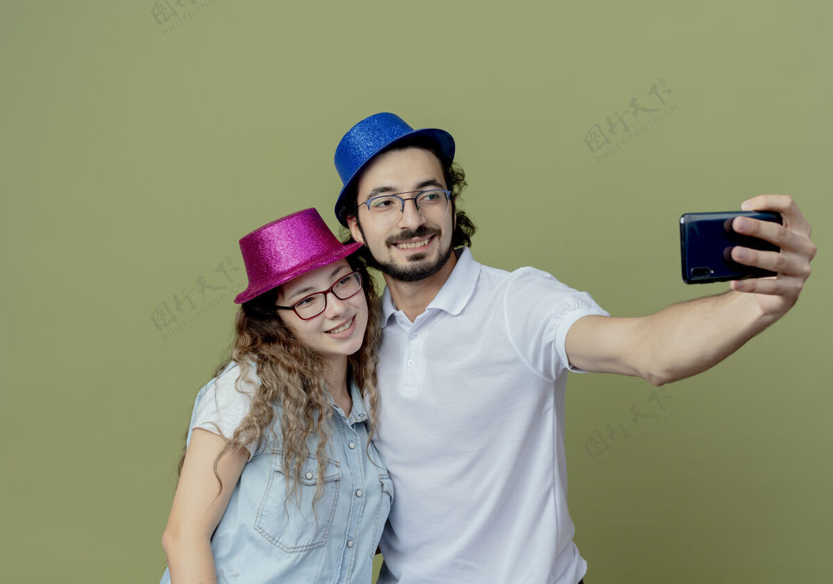 穿着微笑着的年轻夫妇戴着粉色和蓝色的帽子在橄榄绿上自拍橄榄色微笑年轻