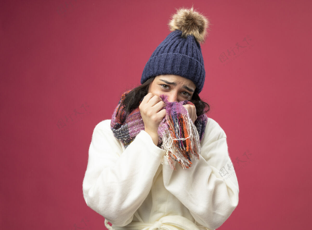 疾病虚弱的生病的年轻女子戴着长袍 戴着冬天的帽子和围巾 用围巾捂着嘴 看着隔离在粉红色墙上的前面围巾人弱者
