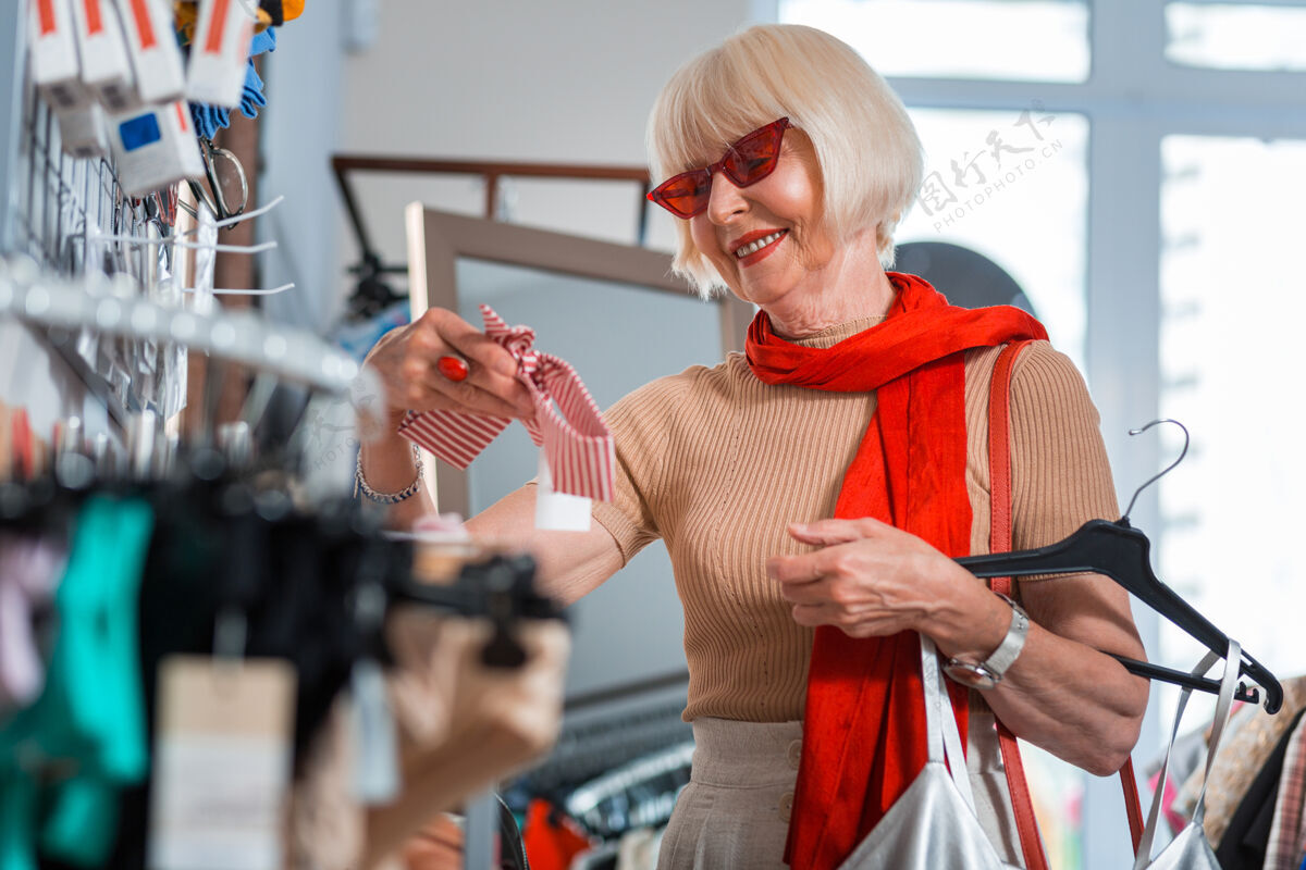 积极衣服的需要部分腰以上的积极的灰色头发的顾客戴着红色太阳镜手持头带 而站在时尚服装店腰围顾客女人