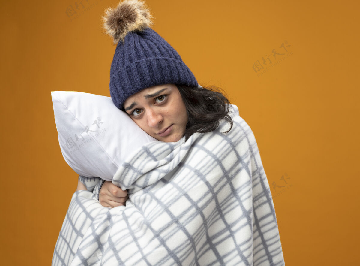 站立体弱多病的年轻女子穿着长袍 戴着格子呢的冬帽 站在侧视图里 抱着枕头 看着橙色墙壁上孤立的前方长袍轮廓冬天