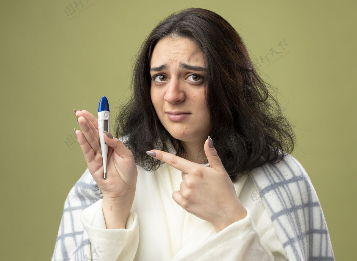 姿势焦急的年轻生病的女人穿着格子布裹着的长袍 对着前面的温度计指着它 看着橄榄绿墙上孤立的前面展示人橄榄