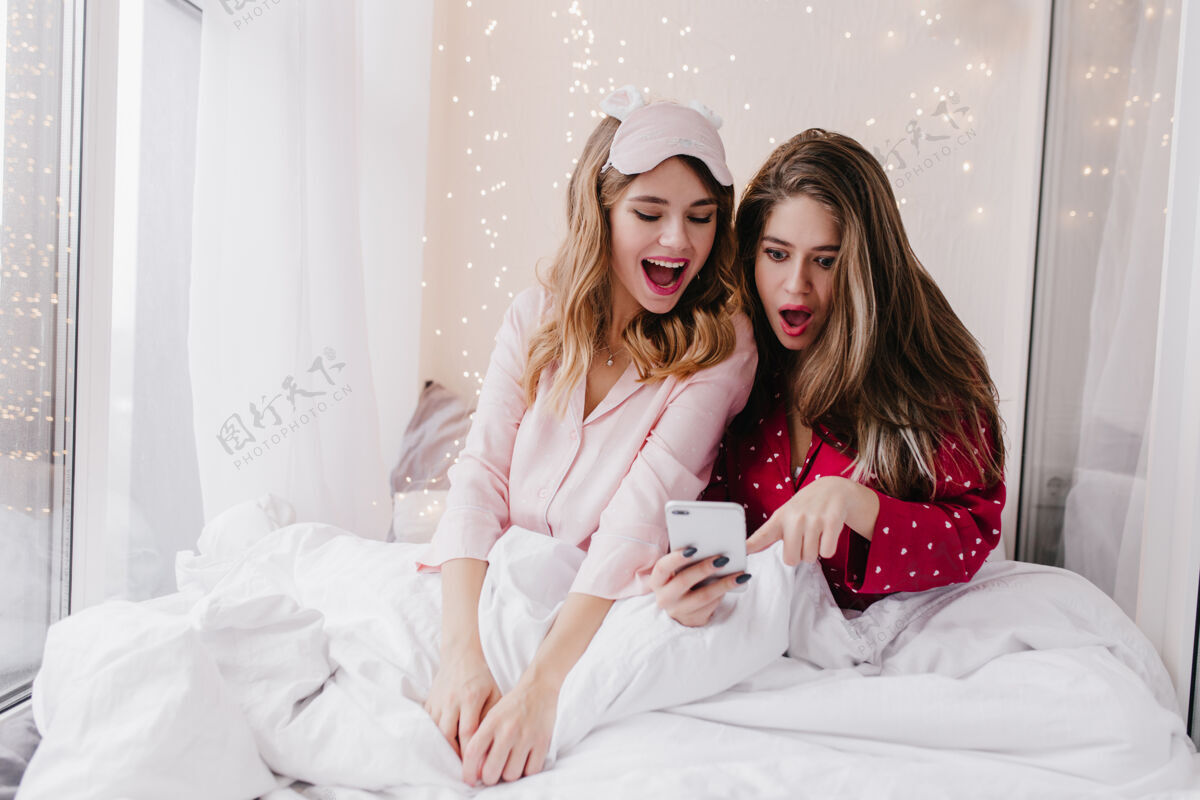 放松震惊的长发女孩坐在床上看着手机屏幕穿着睡衣的女性朋友在光线充足的卧室里摆姿势的照片情绪手机享受
