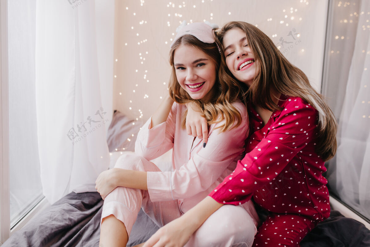 享受美丽的长发女模特拥抱着妹妹 微笑着害羞的卷发女人穿着粉色睡衣在卧室里和朋友拥抱漂亮友谊拥抱