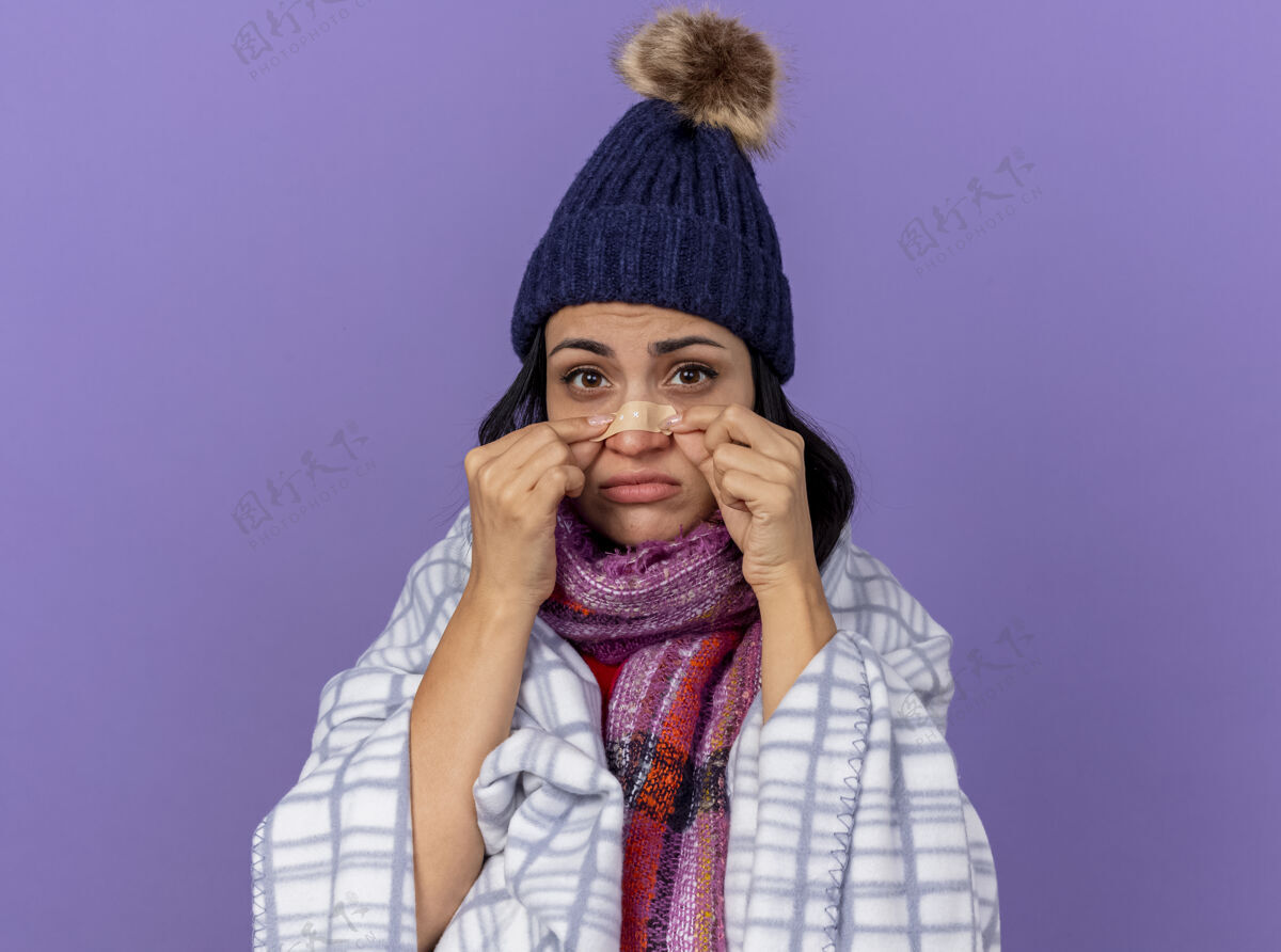 疾病悲伤的生病的年轻女人戴着冬天的帽子 围着格子呢围巾 看着前面贴着膏药的鼻子 隔离在紫色的墙上包裹石膏格子