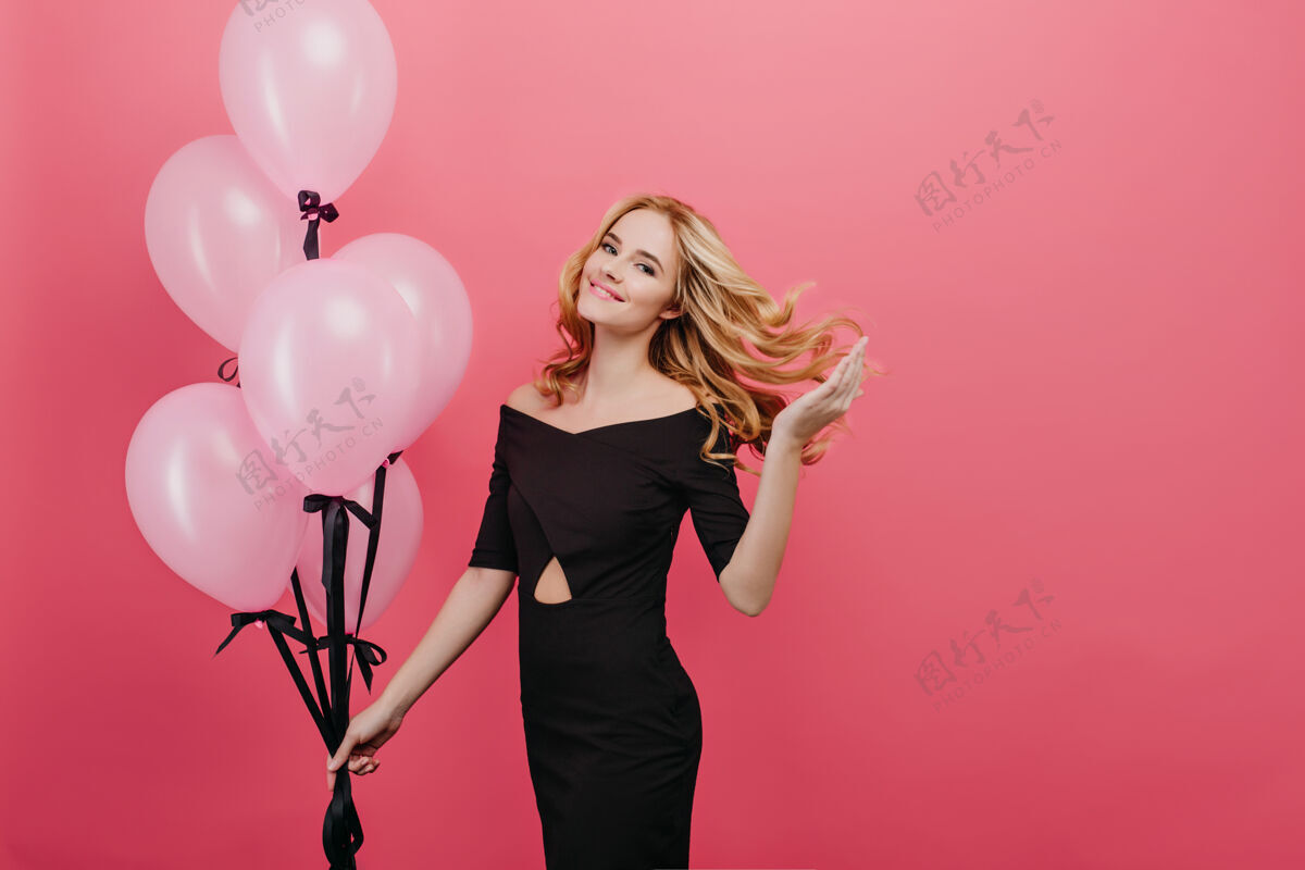气球迷人的苗条女子在与派对气球拍照时玩着卷发优雅的生日女孩身着黑色礼服 在粉色的墙上享受活动并摆出姿势休闲表情粉色气球