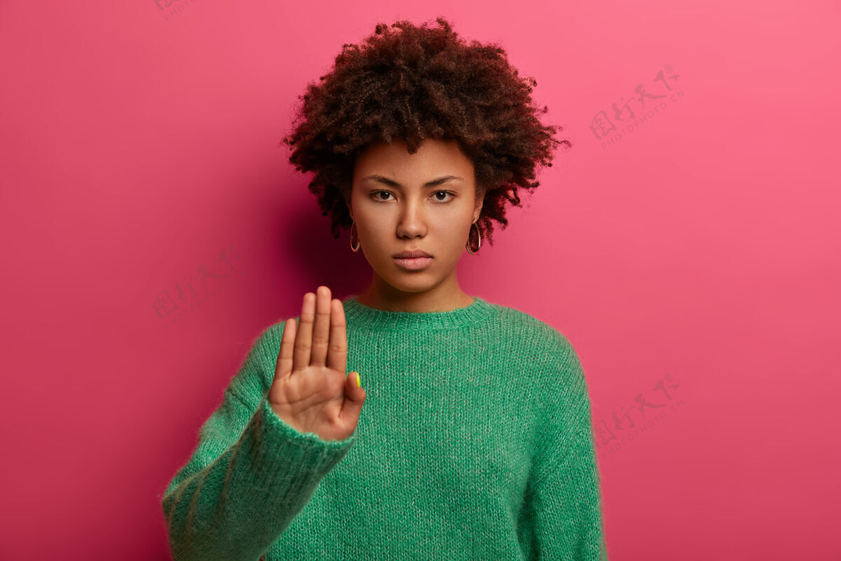 显示年轻严肃的卷发美国黑人妇女用手掌做停车标志 穿绿色毛衣 展示禁止和限制 拒绝什么 模特对着粉色墙壁 说不信心人跳跃