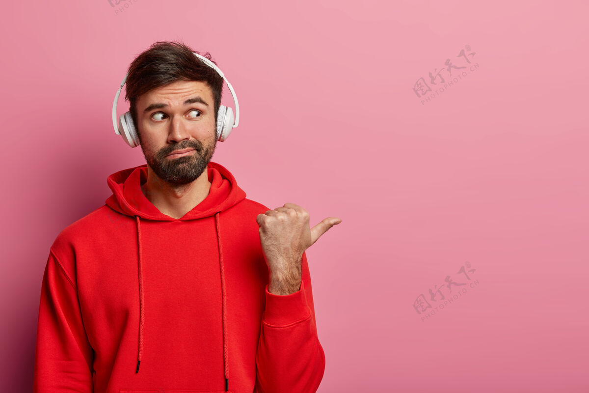帅气好奇的大胡子男人的肖像在空白的右边指着拇指 戴着立体声耳机和红色休闲运动衫 展示了一些有趣的东西 孤立在粉色粉彩墙上站工作时尚
