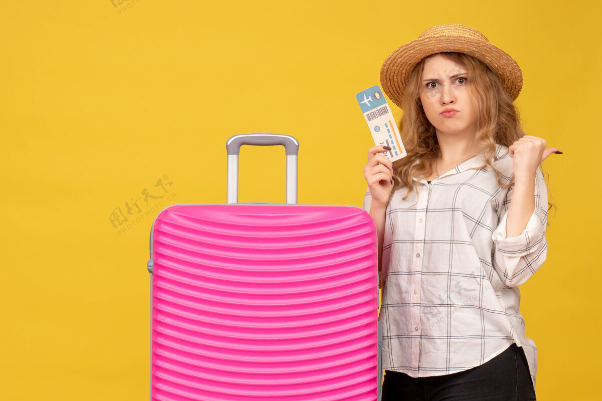 漂亮俯视图：情绪激动的年轻女士戴着帽子 展示着车票 站在她粉红色的包旁 指着后面帽子站购物
